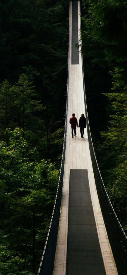 A silhouette of two guests walking in the middle of the iconic Capilano Suspension Bridge, surrounded by breathtaking natural beauty