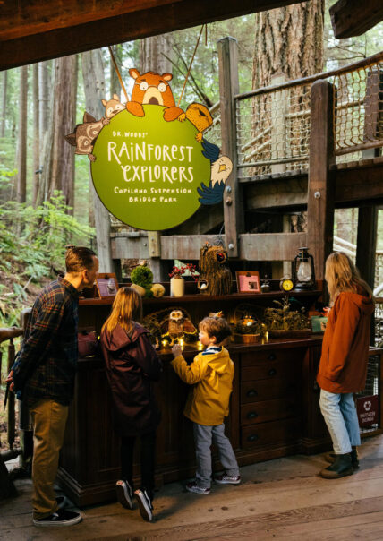 A family gathers in the treehouse of Treetops Adventure, exploring the Kids Rainforest Explorers activity display while the Rainforest Explorers character sign hangs above, adding to the fun and educational experience at Capilano Suspension Bridge Park